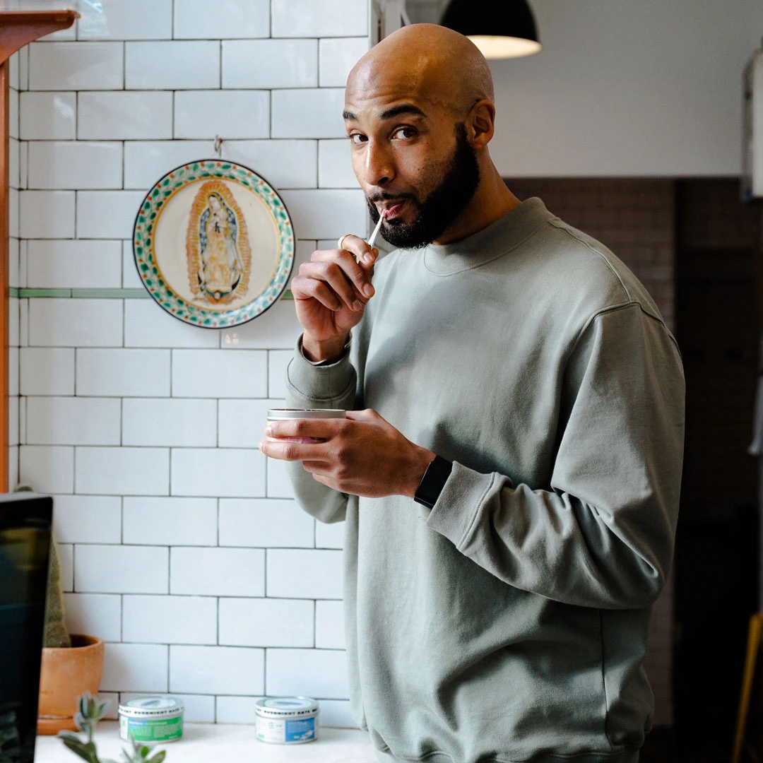 The founder in gray sweatshirt tasting food with a spoon in a kitchen with white tiled walls.
